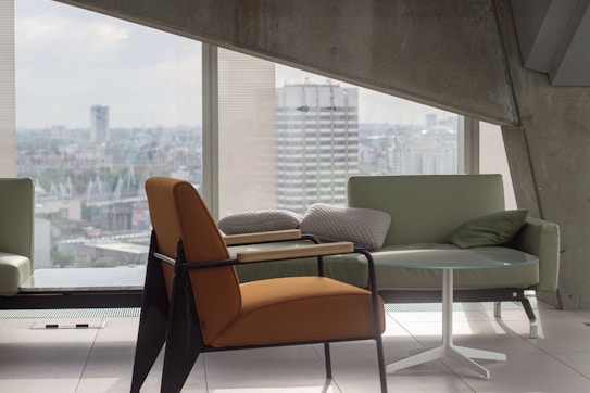 A modern interior scene with a focus on sleek furniture, including an orange armchair, a light green sofa, and a small round glass table. Large windows offer a view of a cityscape, allowing plenty of natural light to flood the space. The architecture features clean lines and concrete elements, contributing to a minimalist aesthetic.