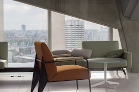 A modern interior scene with a focus on sleek furniture, including an orange armchair, a light green sofa, and a small round glass table. Large windows offer a view of a cityscape, allowing plenty of natural light to flood the space. The architecture features clean lines and concrete elements, contributing to a minimalist aesthetic.