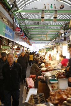 A bustling indoor market corridor with numerous food stalls on one side. Shoppers and vendors are interacting, with some people sitting at counters enjoying meals. The roof is arched with a green frame, and there are signs and advertisements in Korean. The atmosphere is lively and crowded.