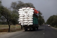 A large truck is overloaded with tightly packed white sacks, secured with ropes, driving along a rural road lined with trees. The sky appears overcast, and there are a few smaller vehicles and bicycles sharing the road in the distance.