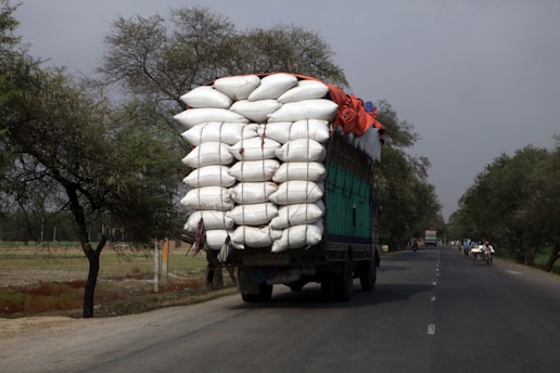 A large truck is overloaded with tightly packed white sacks, secured with ropes, driving along a rural road lined with trees. The sky appears overcast, and there are a few smaller vehicles and bicycles sharing the road in the distance.