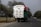 A large truck is overloaded with tightly packed white sacks, secured with ropes, driving along a rural road lined with trees. The sky appears overcast, and there are a few smaller vehicles and bicycles sharing the road in the distance.