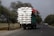 A large truck is overloaded with tightly packed white sacks, secured with ropes, driving along a rural road lined with trees. The sky appears overcast, and there are a few smaller vehicles and bicycles sharing the road in the distance.