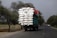 A large truck is overloaded with tightly packed white sacks, secured with ropes, driving along a rural road lined with trees. The sky appears overcast, and there are a few smaller vehicles and bicycles sharing the road in the distance.