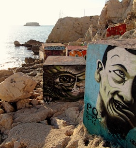 A rocky shoreline with large concrete blocks featuring intricate graffiti art depicting faces and an eye. The ocean stretches into the distance, with a small island visible on the horizon under a bright sky. The scene captures both natural and urban elements, blending the rough texture of the rocks with vivid street art.