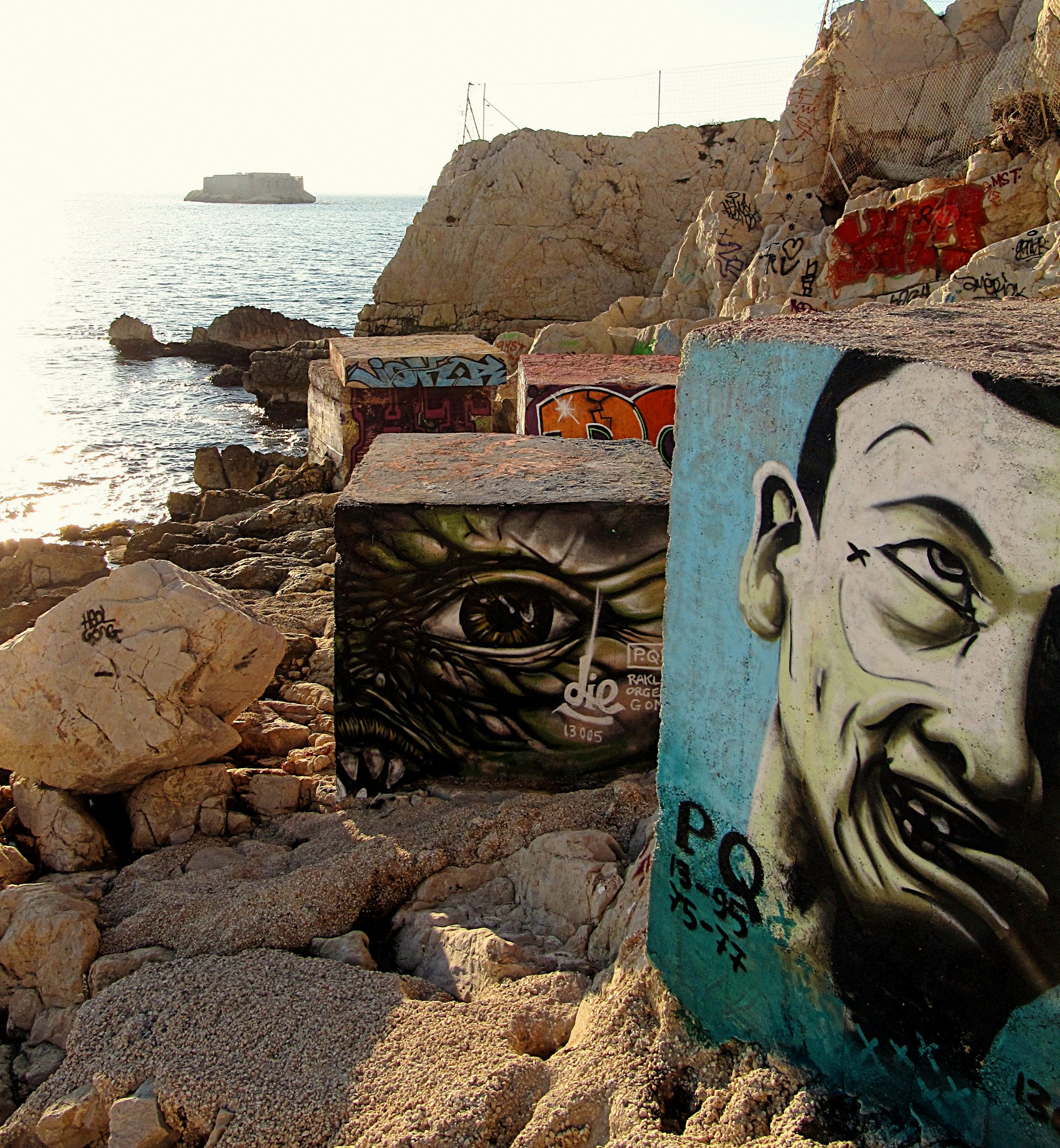 A rocky shoreline with large concrete blocks featuring intricate graffiti art depicting faces and an eye. The ocean stretches into the distance, with a small island visible on the horizon under a bright sky. The scene captures both natural and urban elements, blending the rough texture of the rocks with vivid street art.