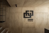 A minimalist interior with warm wooden panels covering the walls and ceiling. A geometric logo and the words 'FRAME HOUSE' are prominently displayed. A black metal fixture is mounted on the left, and a spotlight is suspended from the ceiling, adding to the modern aesthetic. The overall design is sleek and contemporary.
