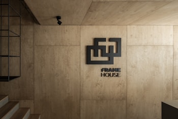 A minimalist interior with warm wooden panels covering the walls and ceiling. A geometric logo and the words 'FRAME HOUSE' are prominently displayed. A black metal fixture is mounted on the left, and a spotlight is suspended from the ceiling, adding to the modern aesthetic. The overall design is sleek and contemporary.