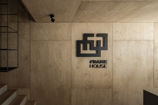 A minimalist interior with warm wooden panels covering the walls and ceiling. A geometric logo and the words 'FRAME HOUSE' are prominently displayed. A black metal fixture is mounted on the left, and a spotlight is suspended from the ceiling, adding to the modern aesthetic. The overall design is sleek and contemporary.