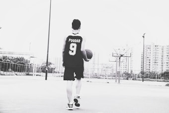 A person wearing a basketball jersey with the name and number 'POODAR 9' is holding a basketball and walking on an outdoor court. The background features tall buildings and multiple basketball hoops. The image is in black and white.