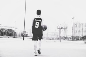 A person wearing a basketball jersey with the name and number 'POODAR 9' is holding a basketball and walking on an outdoor court. The background features tall buildings and multiple basketball hoops. The image is in black and white.