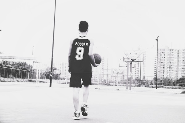 A person wearing a basketball jersey with the name and number 'POODAR 9' is holding a basketball and walking on an outdoor court. The background features tall buildings and multiple basketball hoops. The image is in black and white.