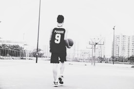 A person wearing a basketball jersey with the name and number 'POODAR 9' is holding a basketball and walking on an outdoor court. The background features tall buildings and multiple basketball hoops. The image is in black and white.