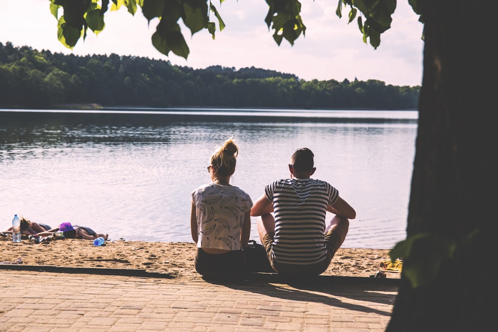 men and woman sitting on pavement facing body of water