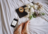 A cozy setting with a smartphone displaying a motivational message, resting on striped fabric. Next to it, a slate board holds a dessert topped with blueberries. A bouquet of lilac flowers with green leaves adds a natural touch, and tattooed feet are visible in the foreground.