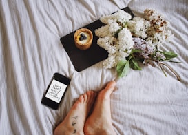 A cozy setting with a smartphone displaying a motivational message, resting on striped fabric. Next to it, a slate board holds a dessert topped with blueberries. A bouquet of lilac flowers with green leaves adds a natural touch, and tattooed feet are visible in the foreground.
