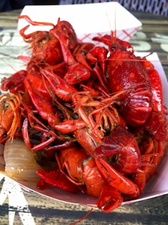 A bowl filled with a pile of bright red crawfish placed on a table. The crawfish are cooked and stacked on top of each other in a white paper container, suggesting a meal ready to be eaten. Some onions are visible underneath the crawfish.