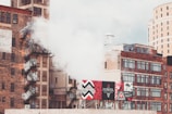 Urban scene with multiple brick buildings, some featuring fire escapes and graffiti. A thick plume of white smoke or steam is rising alongside advertising billboards displaying vibrant colors and designs. The background shows a mix of industrial and more modern architecture under a cloudy sky.