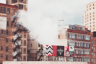 Urban scene with multiple brick buildings, some featuring fire escapes and graffiti. A thick plume of white smoke or steam is rising alongside advertising billboards displaying vibrant colors and designs. The background shows a mix of industrial and more modern architecture under a cloudy sky.