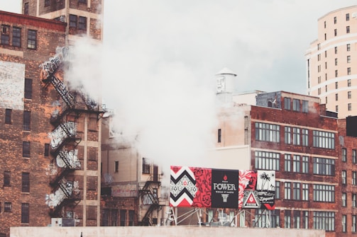 Urban scene with multiple brick buildings, some featuring fire escapes and graffiti. A thick plume of white smoke or steam is rising alongside advertising billboards displaying vibrant colors and designs. The background shows a mix of industrial and more modern architecture under a cloudy sky.