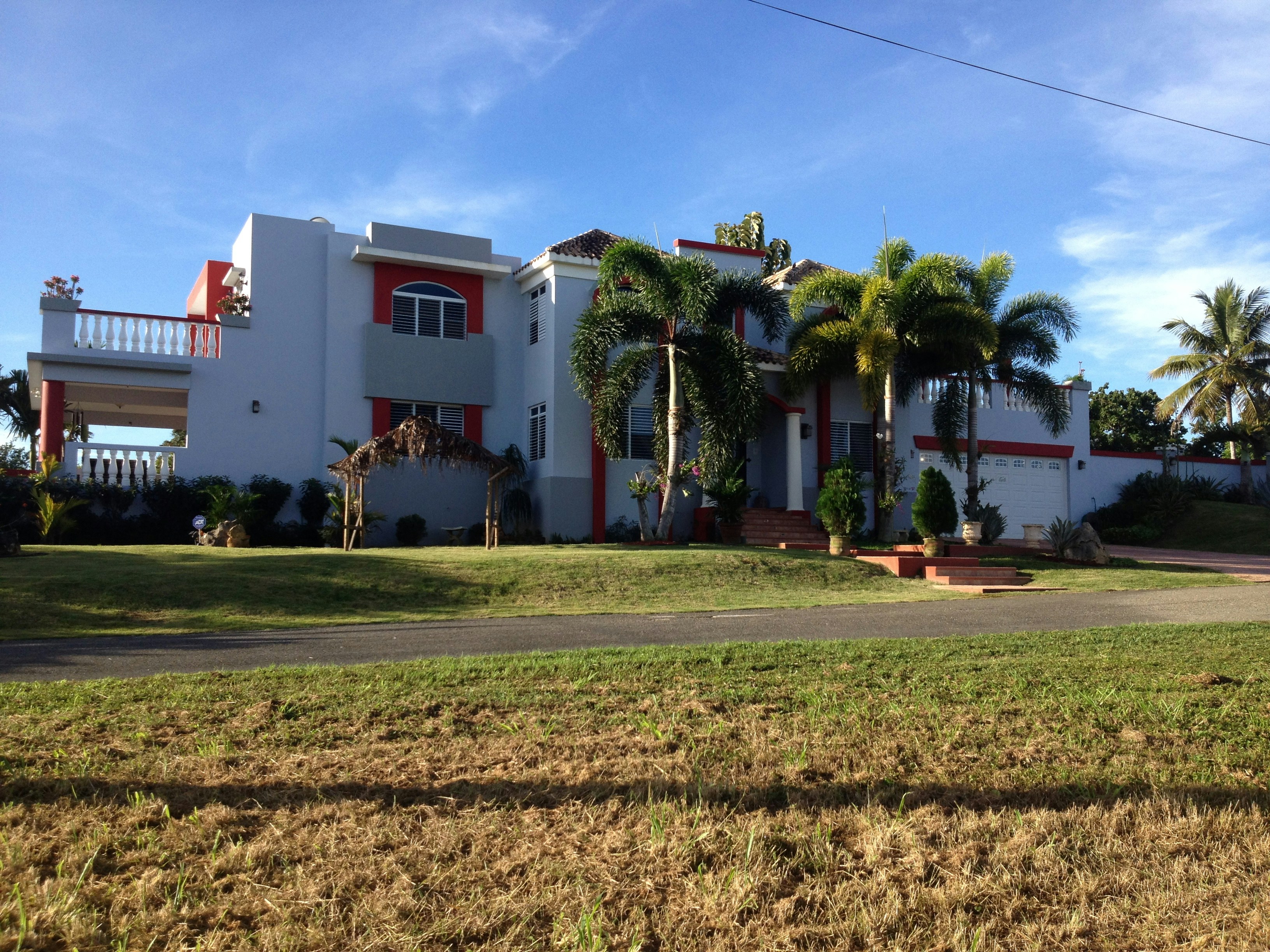 A large, modern house with white walls and red accents, featuring a balcony and several large windows. Palm trees and well-kept landscaping enhance the property, which has a driveway leading to a garage. The sky is clear and the sun casts shadows on the grass lawn.
