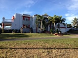 A large, modern house with white walls and red accents, featuring a balcony and several large windows. Palm trees and well-kept landscaping enhance the property, which has a driveway leading to a garage. The sky is clear and the sun casts shadows on the grass lawn.