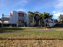 A large, modern house with white walls and red accents, featuring a balcony and several large windows. Palm trees and well-kept landscaping enhance the property, which has a driveway leading to a garage. The sky is clear and the sun casts shadows on the grass lawn.