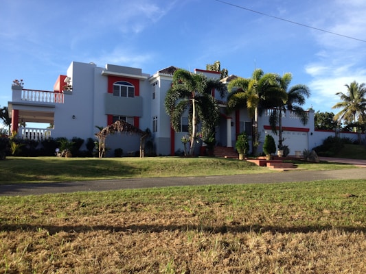 A large, modern house with white walls and red accents, featuring a balcony and several large windows. Palm trees and well-kept landscaping enhance the property, which has a driveway leading to a garage. The sky is clear and the sun casts shadows on the grass lawn.