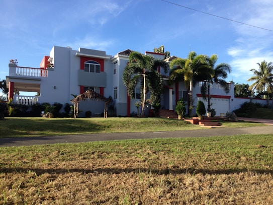 A large, modern house with white walls and red accents, featuring a balcony and several large windows. Palm trees and well-kept landscaping enhance the property, which has a driveway leading to a garage. The sky is clear and the sun casts shadows on the grass lawn.