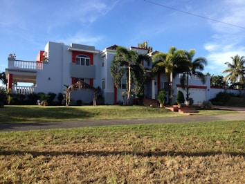A large, modern house with white walls and red accents, featuring a balcony and several large windows. Palm trees and well-kept landscaping enhance the property, which has a driveway leading to a garage. The sky is clear and the sun casts shadows on the grass lawn.