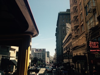 A bustling urban street scene featuring tall buildings on either side. The left side shows part of a historic-looking structure with a strong shadow cast over it. The right side displays a row of older, ornate buildings with fire escapes and commercial signs. Among the signs, one reads 'Sears Fine Food,' indicating a dining establishment. The street below is filled with moving traffic, including cars and what appears to be a cable car partially visible in the foreground. The sky above is clear and blue.