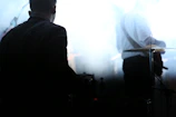 Silhouetted musicians perform in a dimly lit environment, with one person playing the drums and another playing a guitar. A smokey or foggy atmosphere envelops the scene, contributing to a mysterious and moody setting.