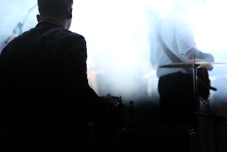 Silhouetted musicians perform in a dimly lit environment, with one person playing the drums and another playing a guitar. A smokey or foggy atmosphere envelops the scene, contributing to a mysterious and moody setting.