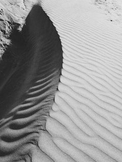 A black and white photograph of sand dunes with distinct ripples and textures. The sand forms elegant patterns, with light and shadow enhancing the undulating shapes. Tracks or trails of footsteps are visible on one side, adding a touch of life to the stark landscape.