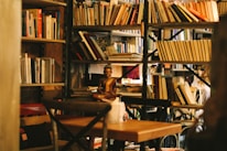 A cozy library or reading area filled with numerous bookshelves. A small statue, possibly of a meditative figure, sits prominently among the books. The environment appears warm and inviting, with a subtle focus on a wooden table and chairs.