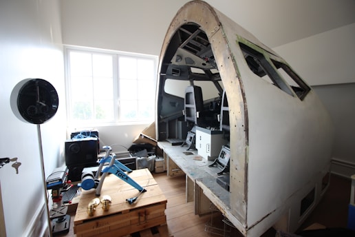 A partial simulation or model of an airplane cockpit positioned in a room. The setup includes two seats and various control panels. The surrounding area contains tools, a wooden crate, electronic devices, and a bicycle trainer.