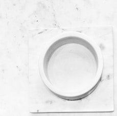 A round, white marble dish sits on a square, white marble slab with a smooth surface. Subtle gray veins and small specks are visible across the marble, enhancing its natural texture.