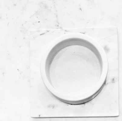 A round, white marble dish sits on a square, white marble slab with a smooth surface. Subtle gray veins and small specks are visible across the marble, enhancing its natural texture.