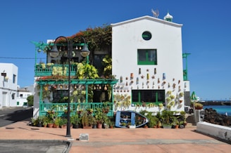 A vibrant, colorful house decorated with numerous potted plants and decorative wall art. The building has a white facade with green accents, including window frames and balcony railings. The house is adorned with various objects such as ceramic art pieces and hanging lights, contributing to its eclectic aesthetic. A path and some street lamps are visible in front, and the house is set against a clear blue sky. Some vegetation grows on the building's balconies and facade.
