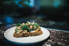 A plate holds two slices of rustic bread topped with a mix of chopped vegetables, herbs, and seeds. The focus on the dish suggests a culinary presentation, perhaps in a dimly lit setting such as a restaurant or home kitchen. Fresh cilantro leaves add a touch of green, complementing the creamy and crunchy textures visible in the topping.