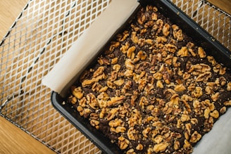 A baking pan filled with a dark, baked dessert topped with abundant pieces of walnuts rests on a cooling rack. The dessert is lined with parchment paper, which extends out of the pan, adding a rustic touch.