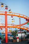 A vibrant amusement park scene with a large Ferris wheel in the background featuring colorful gondolas. An orange roller coaster track winds through the scene. Palm trees and various structures are present below the rides, suggesting a coastal location.