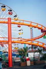 A vibrant amusement park scene with a large Ferris wheel in the background featuring colorful gondolas. An orange roller coaster track winds through the scene. Palm trees and various structures are present below the rides, suggesting a coastal location.
