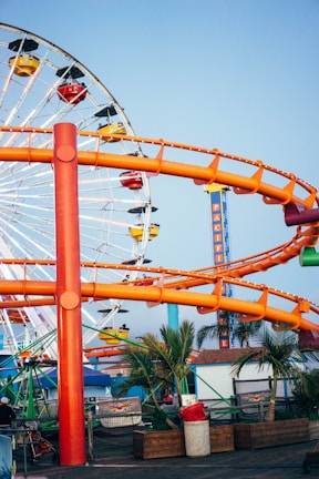 A vibrant amusement park scene with a large Ferris wheel in the background featuring colorful gondolas. An orange roller coaster track winds through the scene. Palm trees and various structures are present below the rides, suggesting a coastal location.