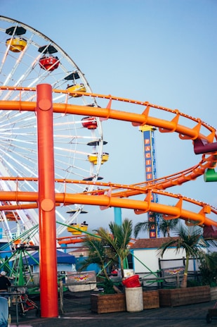A vibrant amusement park scene with a large Ferris wheel in the background featuring colorful gondolas. An orange roller coaster track winds through the scene. Palm trees and various structures are present below the rides, suggesting a coastal location.