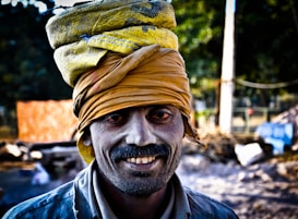 A person wearing a yellow turban and a blue shirt smiles directly at the camera. The background includes blurred natural elements and possibly a construction site with some indistinct objects and structures.