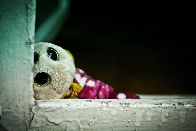 A plush toy with large, shiny eyes peeks out from behind a rough, textured surface. The toy has a white face and is wrapped in a pink polka-dotted fabric. The background is dark, creating a contrast with the brightly colored toy.