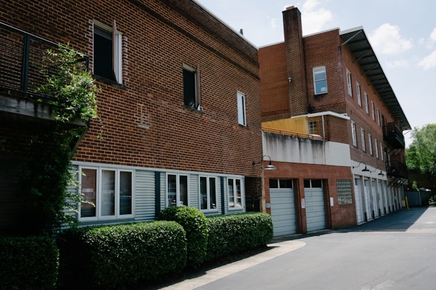 A red brick building with multiple floors, featuring a series of rectangular windows and a row of garage doors on the ground level. The building is surrounded by neatly trimmed green hedges and a small, paved courtyard. There are metal railings along some of the windows and a few outdoor lights mounted on the wall.