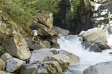 A person is navigating across large boulders near a fast-flowing river within a rocky gorge. The area is surrounded by lush green trees and undergrowth. The river cascades over the rocks creating rapids, and there is a bridge visible in the background above the ravine.