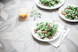 A table setting with plates of fresh salad containing leafy greens and roasted vegetables. A glass of orange juice is placed nearby, and a fork rests on a napkin. The table has a decorative geometric pattern, and a sprig of lavender lies beside the juice.